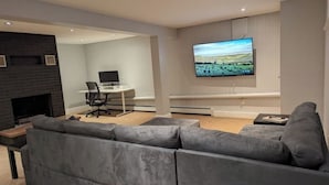 Ein modernes Wohnzimmer mit grauem Sofa, einem Wand-Fernseher, einem Kamin und einem Schreibtisch mit Stuhl.