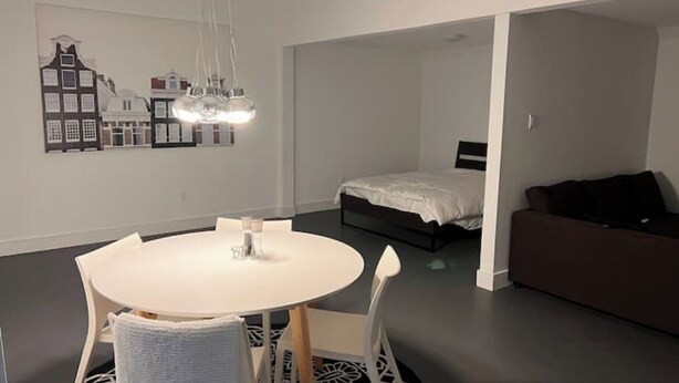 Une pièce moderne et minimaliste, avec une table de salle à manger ronde, des chaises blanches, un lit recouvert d’une couette blanche et un canapé marron.