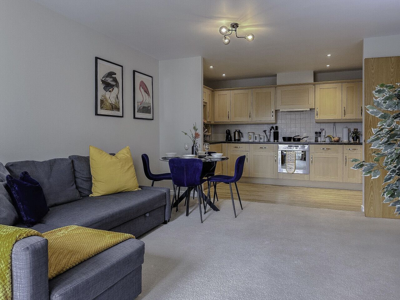A modern living room with a grey sofa, blue chairs, and a small dining table. A kitchen with wooden cabinets and a built-in oven is visible in the background.