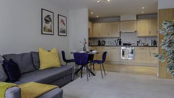 A modern living room with a grey sofa, blue chairs, and a small dining table. A kitchen with wooden cabinets and a built-in oven is visible in the background.