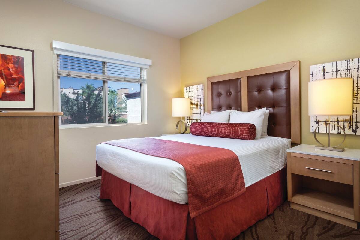 Habitación de hotel con una cama grande, un escritorio con computadora, una ventana con persianas y obras de arte en las paredes.