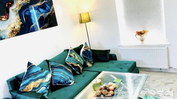 Una sala de estar con un sofá verde, almohadones decorativos, una mesa de centro de vidrio con adornos, una lámpara y un cuadro en la pared.