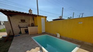 Una piscina con cubierta de baldosas, una pequeña construcción de ladrillo con chimenea y un muro amarillo.