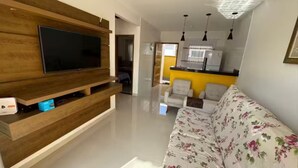 Una sala moderna con una televisión de pantalla plana montada en una pared de madera, un sofá con tapicería floral y una cocina visible al fondo.
