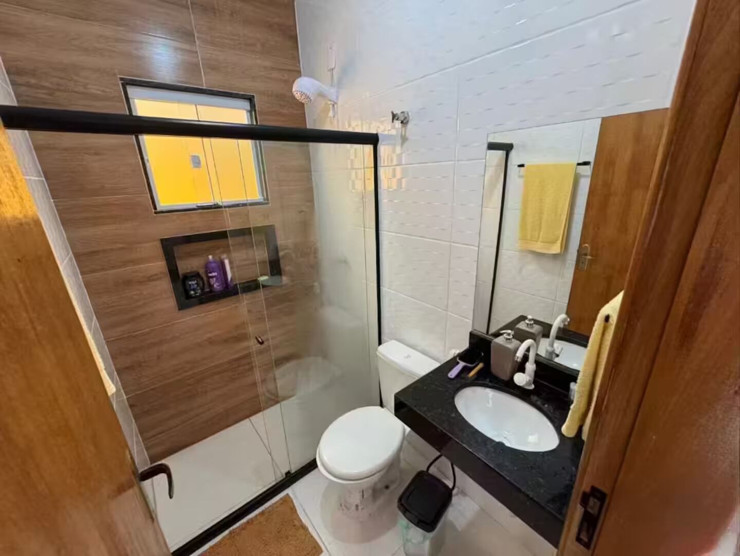 Ein modernes Badezimmer mit Duschabtrennung aus Glas, einer weißen Toilette, einem schwarzen Waschbecken mit Spiegel und einem gelben Handtuch an der Wand.