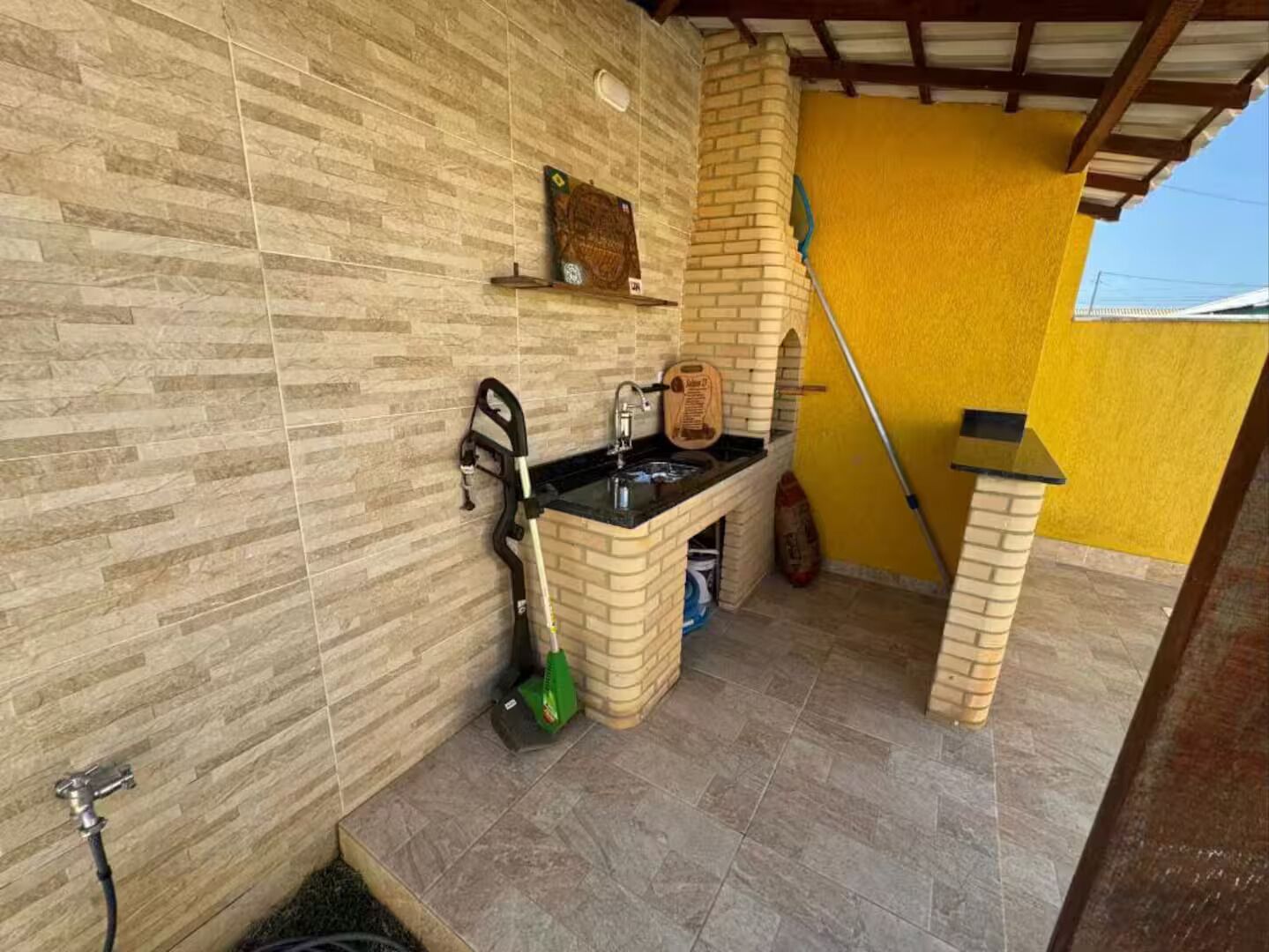A small outdoor kitchen area with a sink, a broom, and a mop leaning against the wall.