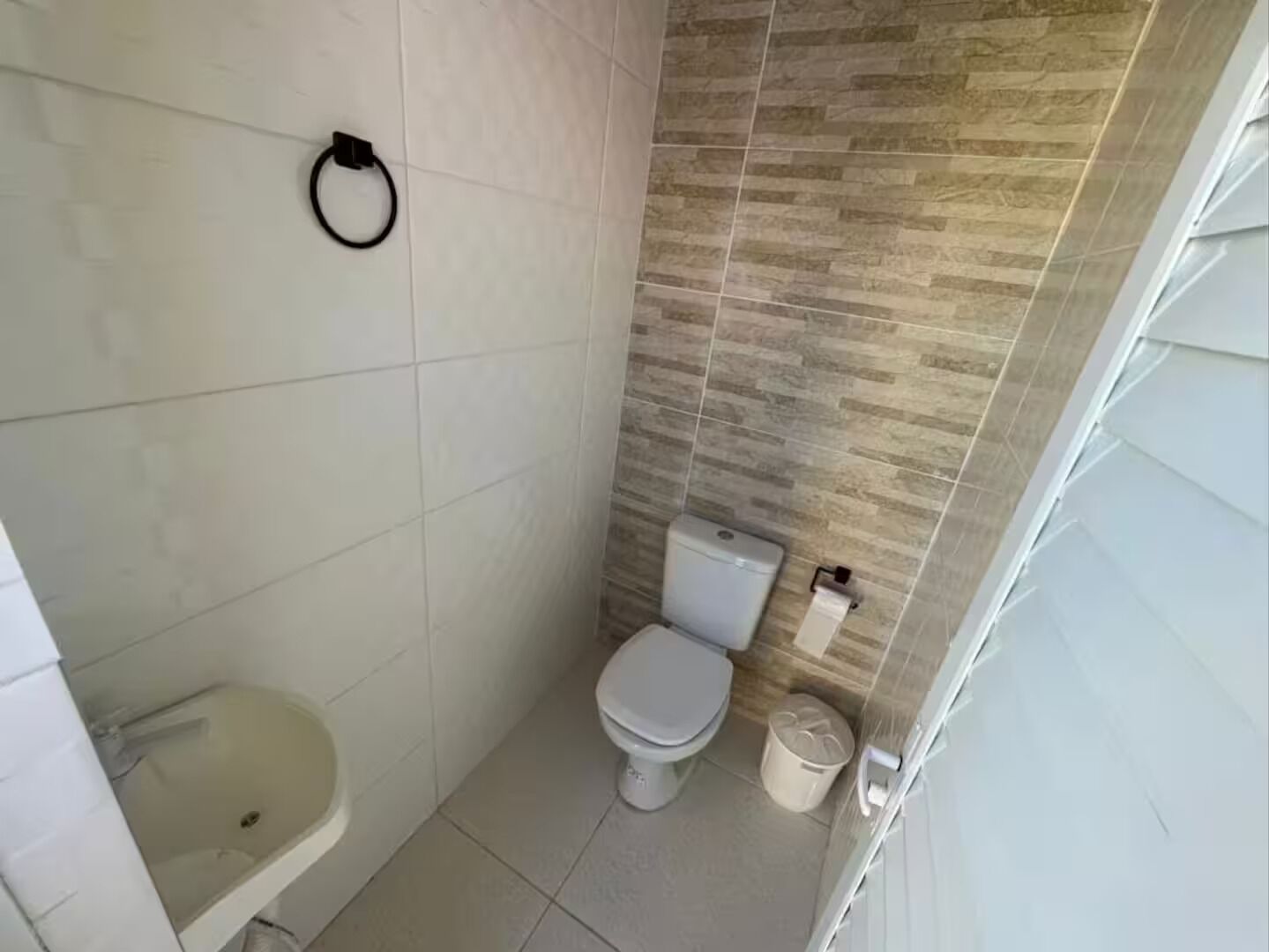 Ein kleines, modernes Badezimmer mit weißen Fliesen, Toilette, Waschbecken und Mülleimer.