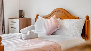 Een netjes opgemaakt bed met witte en roze beddengoed, een houten hoofdbord en een nachtkastje met een lamp en een klein doosje.