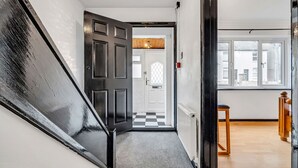 Een gang met een zwarte deur die leidt naar een kamer met een zwart-wit geruite vloer en een raam.