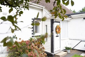 A white house with a black door, a wreath on the door, and a small garden with pink flowers.