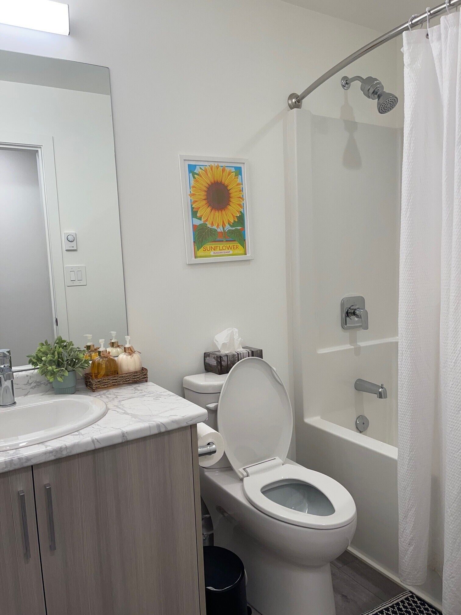 A bathroom with a toilet, sink, shower, and a framed sunflower artwork.