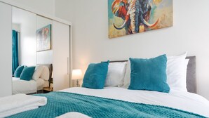 Une chambre d’hôtel avec un grand lit, une literie aux couleurs turquoise et blanche, un tableau représentant un éléphant et un miroir qui reflète la pièce.