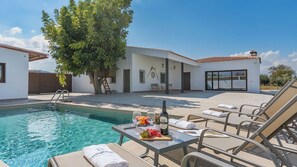 Un espace piscine avec des chaises longues, une table sur laquelle se trouvent une bouteille et des fruits, et un bâtiment doté d’un toit en tuiles.