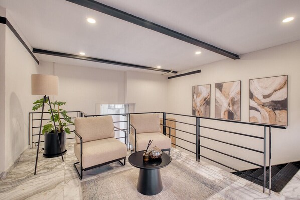 A modern hotel room with a marble floor, two beige armchairs, a coffee table, a floor lamp, and abstract wall art.