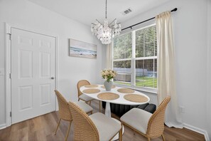 A dining area with a round table, wicker chairs, a vase with flowers, and a view through large windows. - NEW! Game Room~HotTub~Fire Pit~.8mi to Old Town (Bluffton)