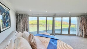 A bedroom with a large bed, a painting on the wall, and a view of the countryside through large windows. - Whitewater, with expansive sea views and hot tub (Westward Ho!)