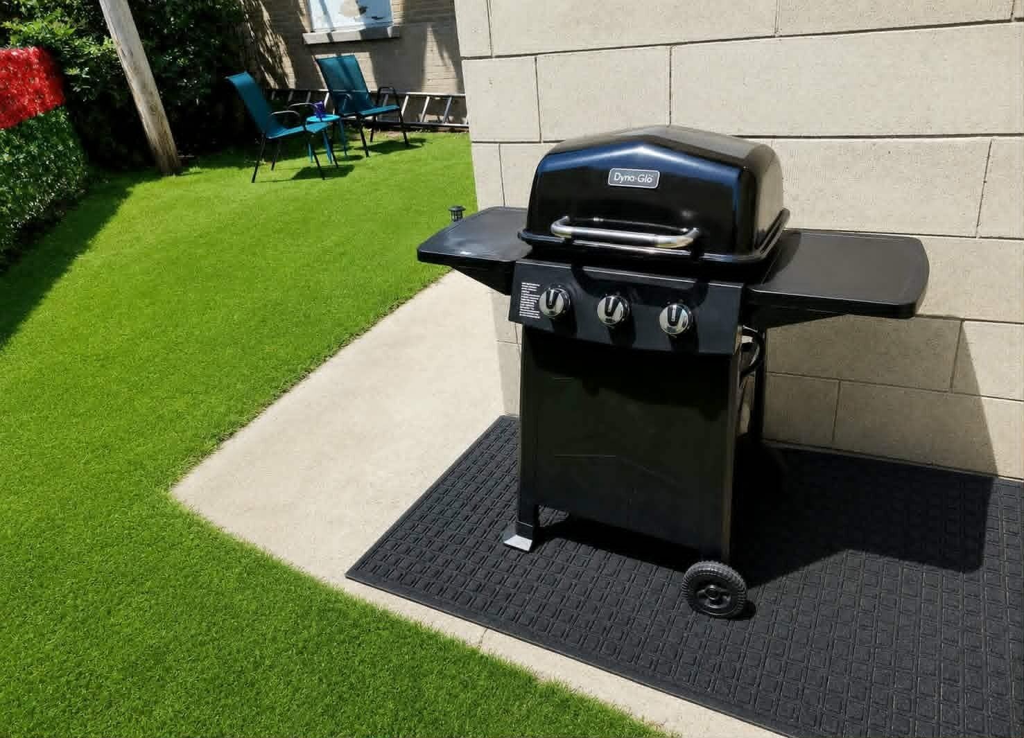 Una parrilla exterior negra con tres perillas de control y una etiqueta con el nombre de la marca, ubicada sobre una superficie pavimentada con un pequeño escalón.
