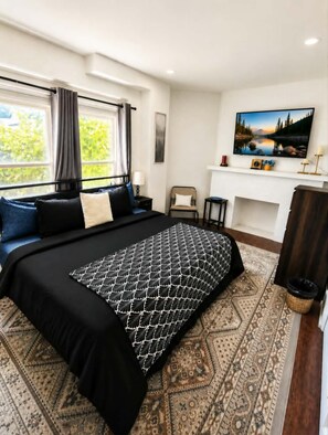 A hotel room with a large bed, a television, a fireplace, and a view of greenery outside. - The Victorian House - 4BR Downstairs Unit • Pet-Friendly • Outdoor Patio & Grill (Buffalo)