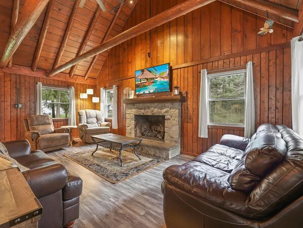 A log cabin interior with a stone fireplace, leather furniture, and a painting above the mantel.