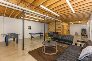 A spacious indoor area with a high ceiling, exposed wooden beams, a foosball table, a pool table, and a comfortable seating area with a sofa and a coffee table.