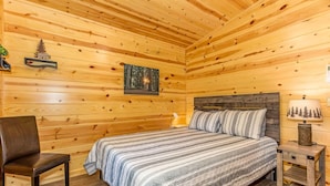 A wooden cabin bedroom with a bed, a chair, a nightstand, and a wall-mounted lamp.