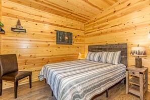 A wooden cabin bedroom with a bed, a chair, a nightstand, and a wall-mounted lamp.