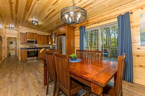 A wooden cabin interior with a dining table, chairs, a kitchen with appliances, and a large window. - Hocking Hills Cabin w/ Hot Tub- near Old Mans Cave (Logan)