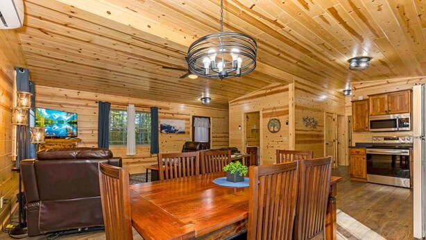 A wooden cabin interior with a dining table, chairs, a sofa, a TV, and a kitchen area.