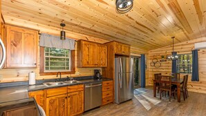 A wooden cabin interior with a kitchen, dining area, and a large window.