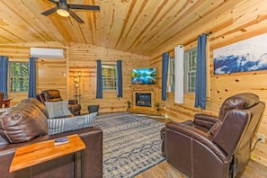 A cozy log cabin interior with leather sofas, a fireplace, a television, and a large window with curtains.