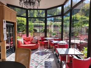 A sunlit café with red furniture, large windows, and a view of a lush garden.