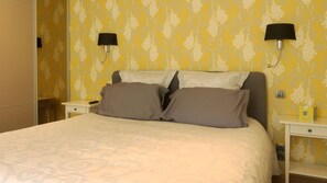 Una cama bien hecha con dos almohadas, una mesita de noche con una lámpara y una pared con estampado floral.
