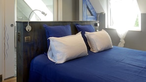A bedroom with a wooden headboard, a large window with curtains, and a bed with blue bedding and white pillows.