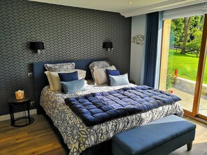 A modern bedroom with a patterned bedspread, a large window with a view of a garden, and a dark blue accent wall.
