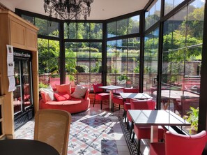 A sunlit café with red furniture, large windows, and a view of a lush garden.