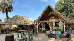A beachside resort with thatched-roof structures, outdoor seating, and a sandy ground. - Curacao,Piscadera Bay, next to Marriott, Very luxuriously furnished Apartment (Willemstad)