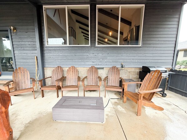 Un espacio al aire libre cubierto con seis sillas Adirondack y una mesa rectangular, una parrilla de barbacoa y un muro de piedra.