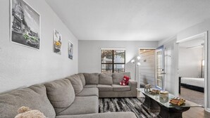 A modern living room with a sectional sofa, a glass coffee table, and a large window with blinds. - THE LITTLE HAVANA AT RIVERSIDE #11 (Austin)