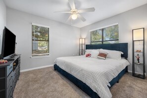 Un dormitorio con cama, ventilador de techo, televisor y dos ventanas con persianas.