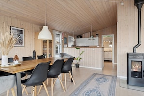 A modern kitchen with wooden cabinets, a dining area with a wooden table and black chairs, a large pendant light, and a wood-burning stove. - Active coastal holiday in cozy cottage (Slagelse)