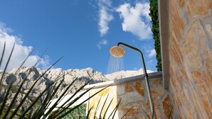 A showerhead with water flowing into a stone wall, set against a backdrop of a clear blue sky and distant mountains.