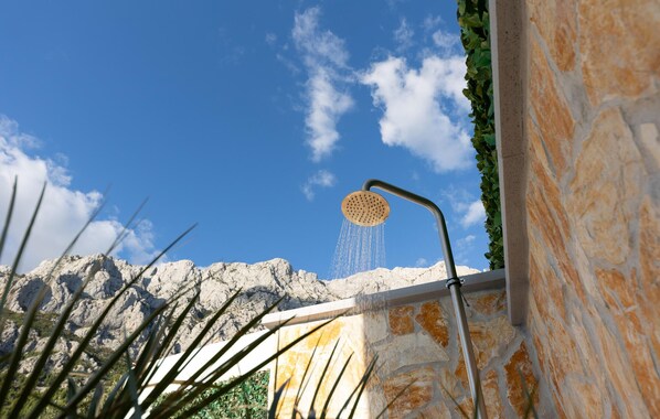A showerhead with water flowing into a stone wall, set against a backdrop of a clear blue sky and distant mountains.