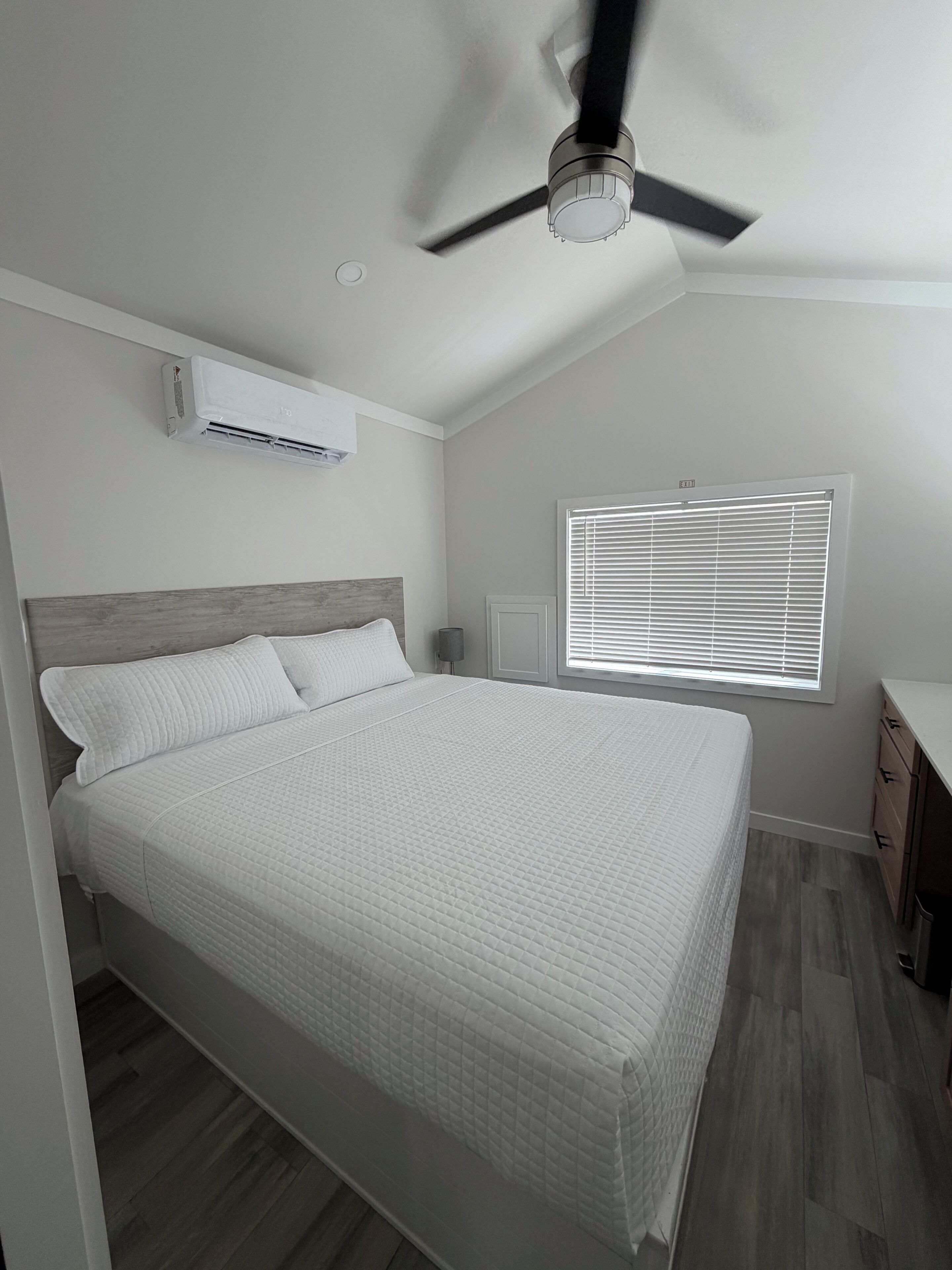 Una camera da letto con un letto, un condizionatore, una ventola a soffitto, una finestra con persiane e un comodino.