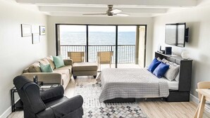 Een slaapkamer met een bed, een bank, een stoel, een bureau en een televisie. Er is een balkon met uitzicht op zee.