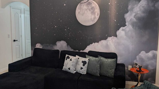 A living room with a black sectional sofa, a patterned rug, and a wall mural of a moon and clouds. - Magical World (Davenport)