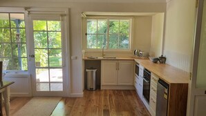 A kitchen with wooden flooring, white cabinets, a window with a view of greenery, and a trash can. - Magnolia Cottage- wineries, main street, station (Croydon North)