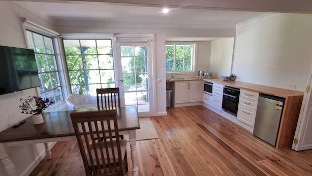 A modern kitchen with wooden flooring, white cabinetry, and a dining area with a wooden table and chairs. - Magnolia Cottage- wineries, main street, station (Croydon North)