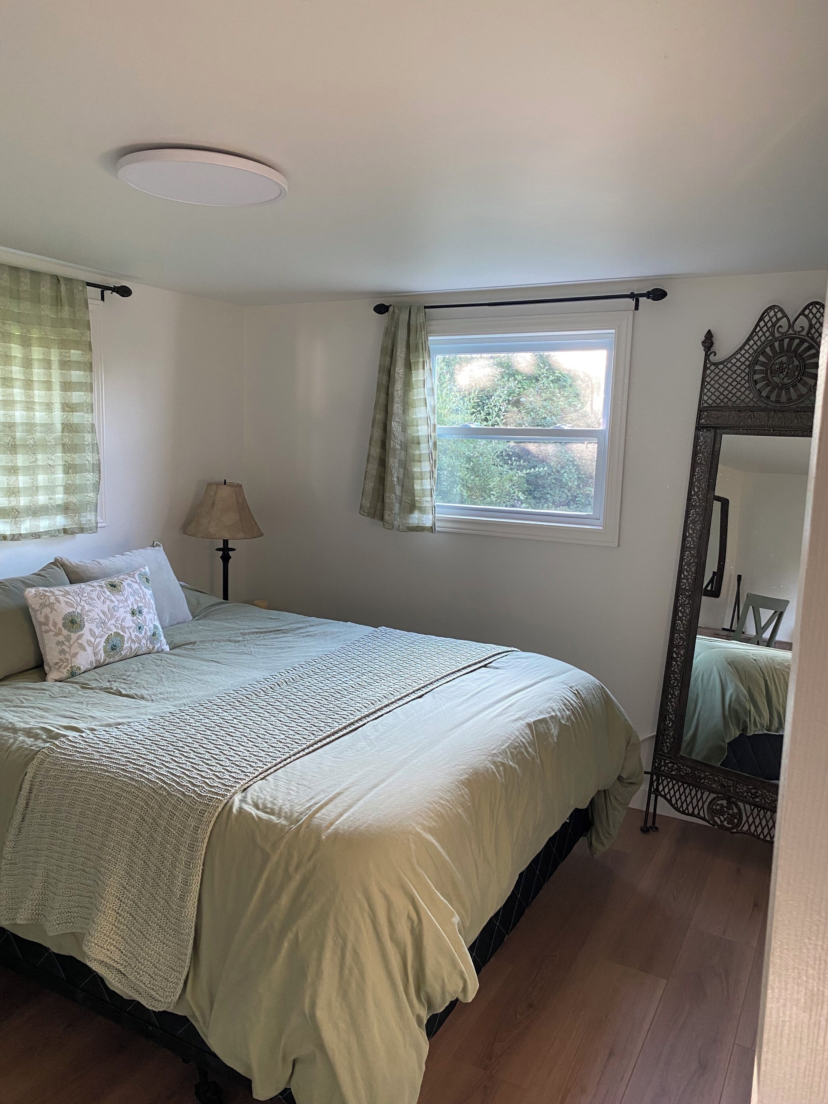 A bedroom with a bed, a window with curtains, a lamp, and a mirror.