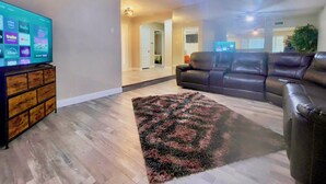 A living room with a flat-screen TV displaying streaming services, a brown leather sofa, a patterned rug, and a ceiling fan.
