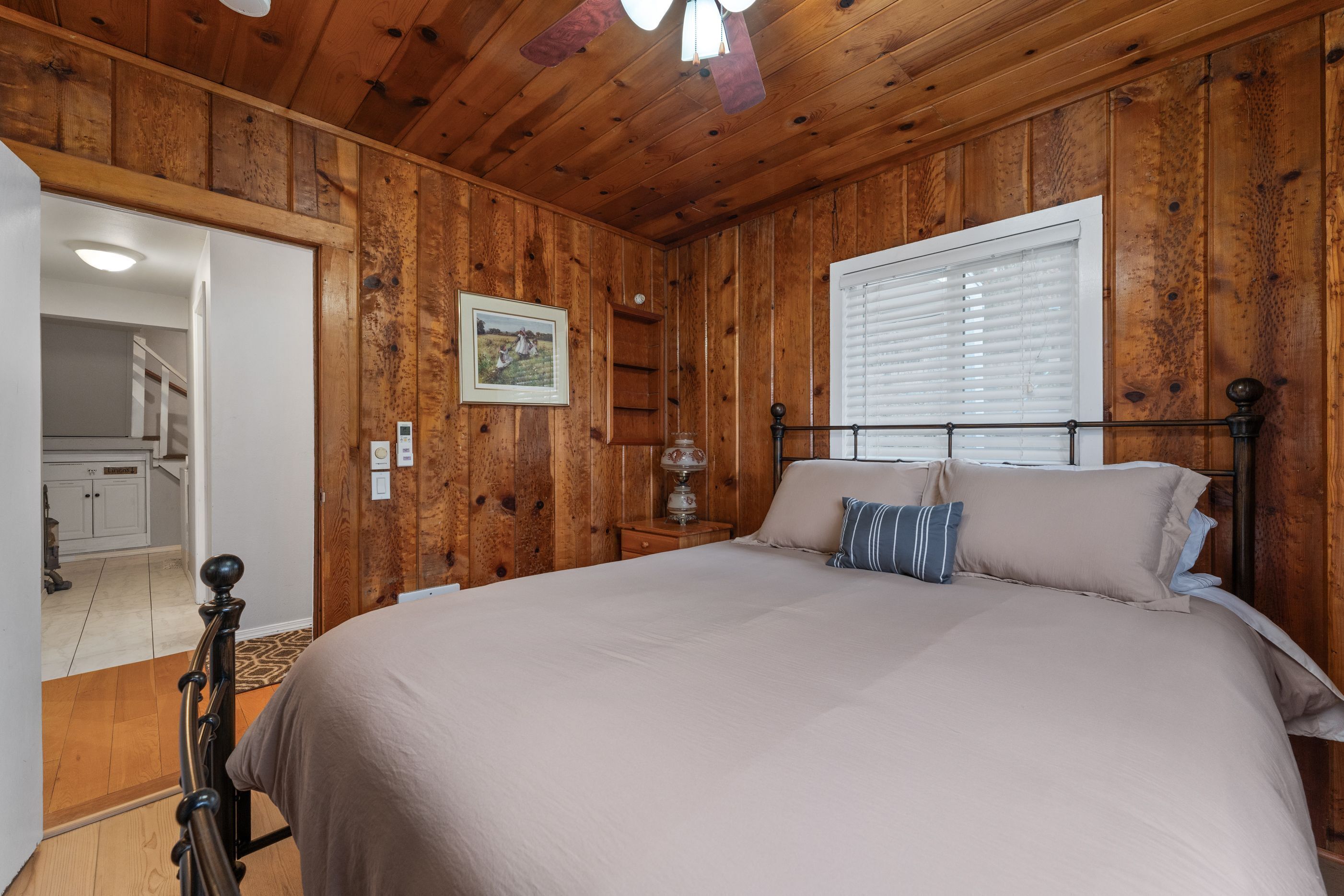 A bedroom with wooden paneling, a bed with beige bedding, a nightstand with a lamp, and a window with blinds.